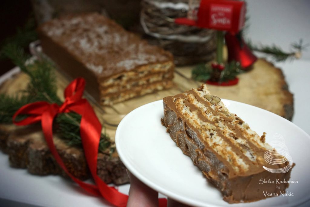 Mamina torta s datuljama - šnita torte servirana na bijelom četvrtastom tanjuru, u pozadini drvena podloga sa tortom i blagdanskim detaljima u zeleno-crvenim bojama